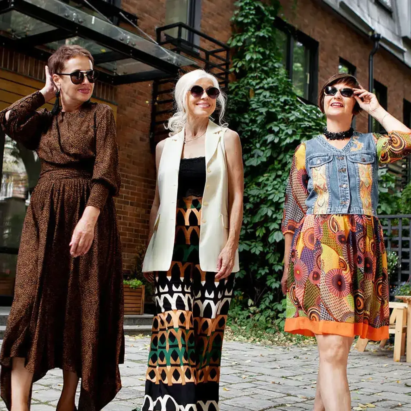 Three stylish women wearing sunglasses walking together along a city street on a sunny day.