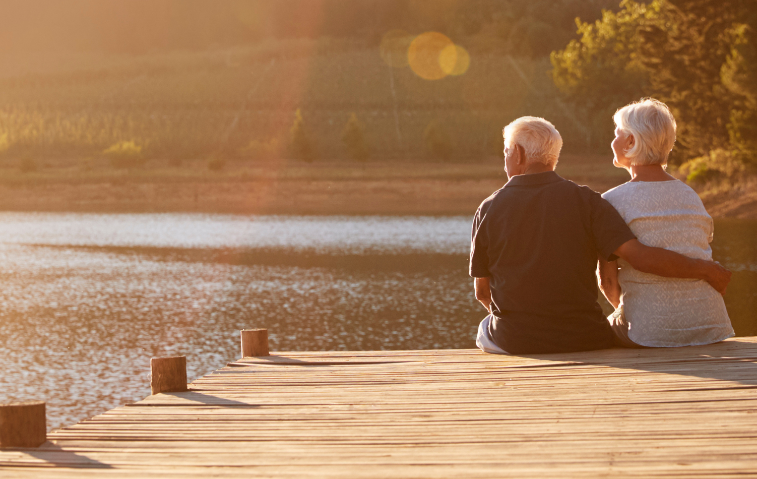 A senior couple sat on the end of a boardwalk looking out over a lake in the sunset