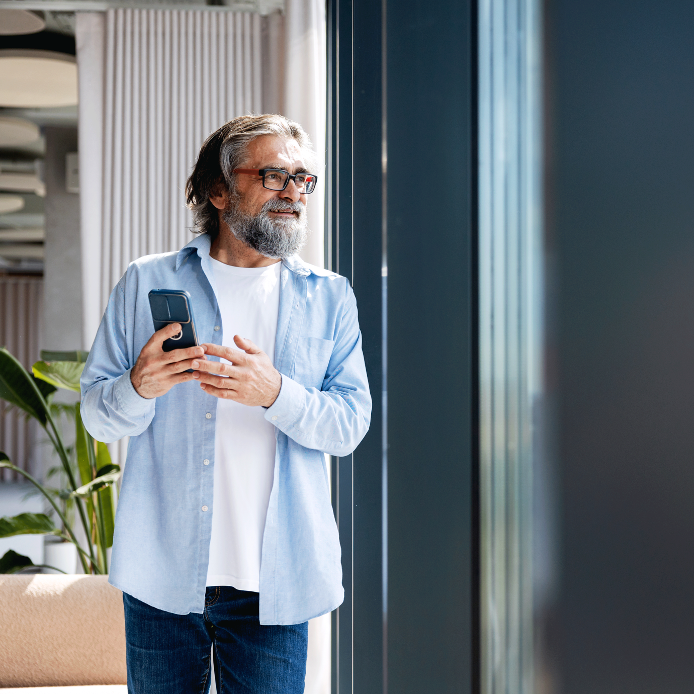 Smiling senior man with grey hair and a beard holding a smartphone while standing by a window.