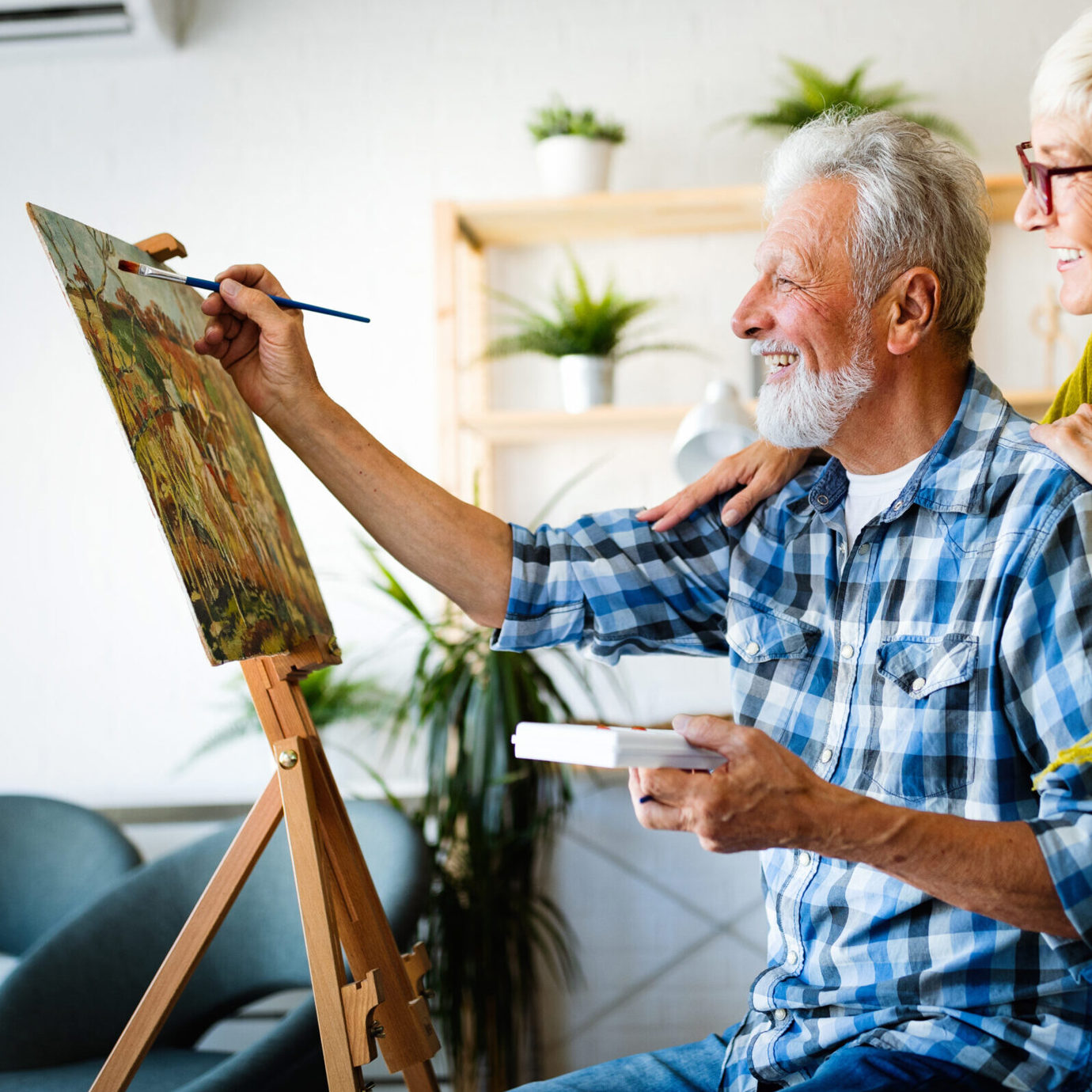 Senior man painting on a canvas while a smiling woman watches and supports him at home.