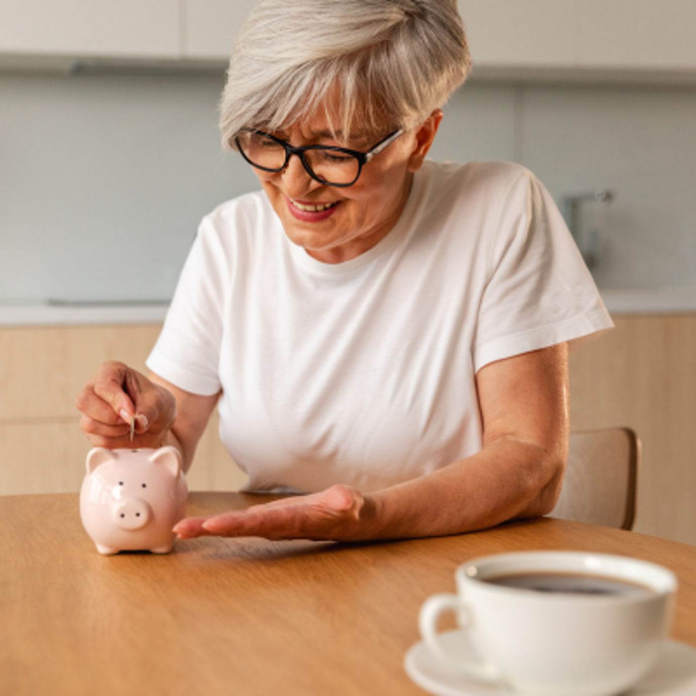 A woman wearing glasses is sat at her kitchen table putting a coin in to her piggy bank