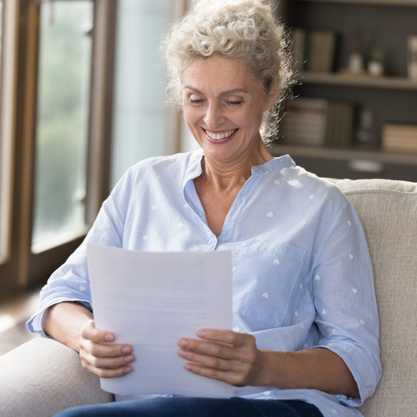Smiling woman with curly grey hair sitting on a sofa at home reading a document.