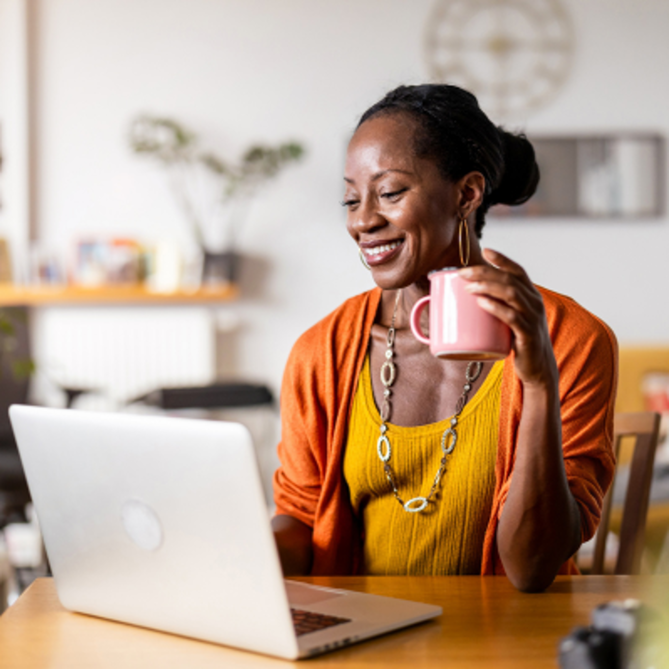 Smiling woman in an orange cardigan holding a pink mug while using a laptop at home.