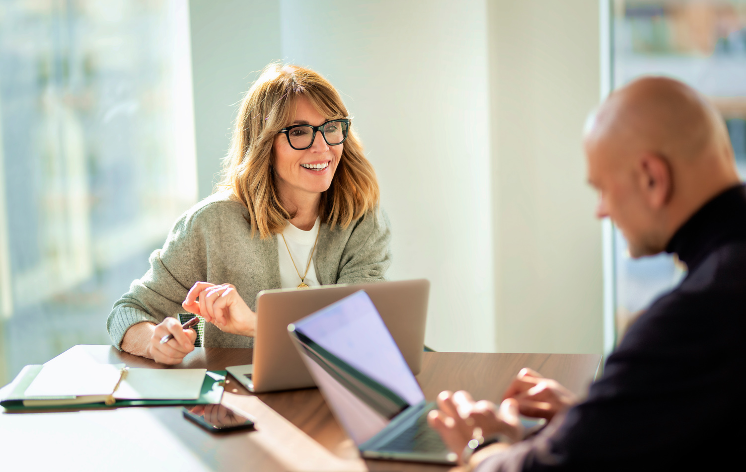 A woman and a man in an office completing work and having a discussion. The woman is centre and has a blue to purple gradient hollow circle encompassing her