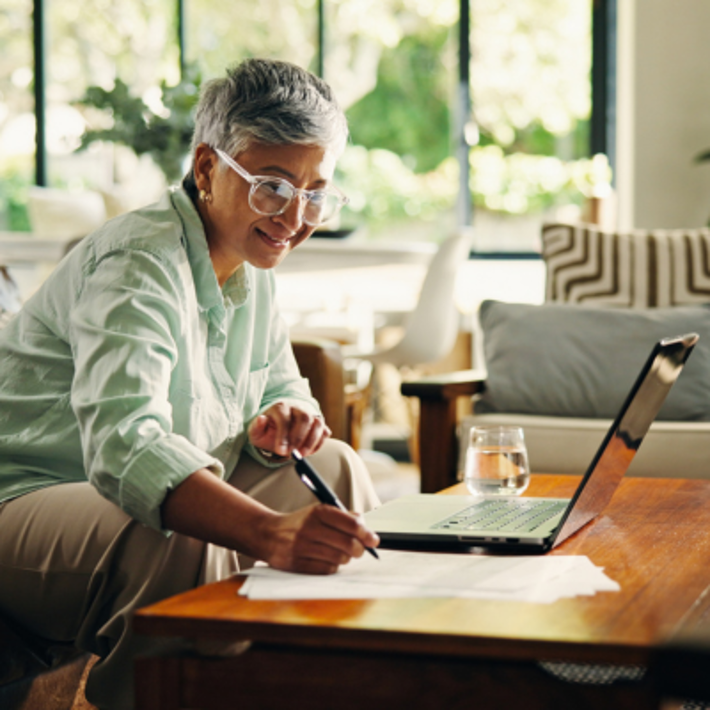 Senior woman wearing glasses reviewing paperwork beside a laptop at home.